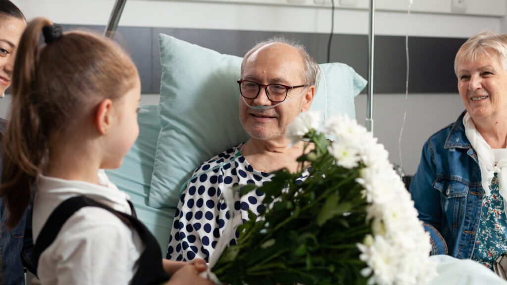 An older patient in hospital, happy, surrounded by family and receiving flowers from his granddaughter